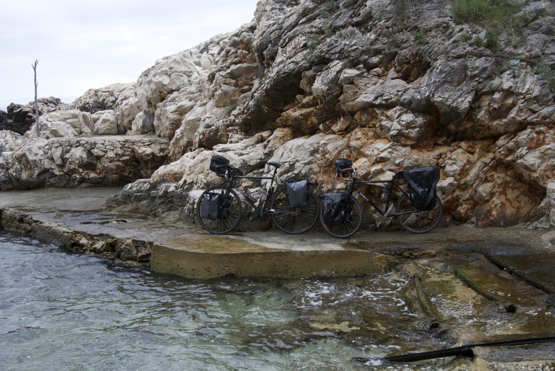 Left side view of two black Surly bikes with gear, leaning on a rock wall base, on a flat stone bank next to water