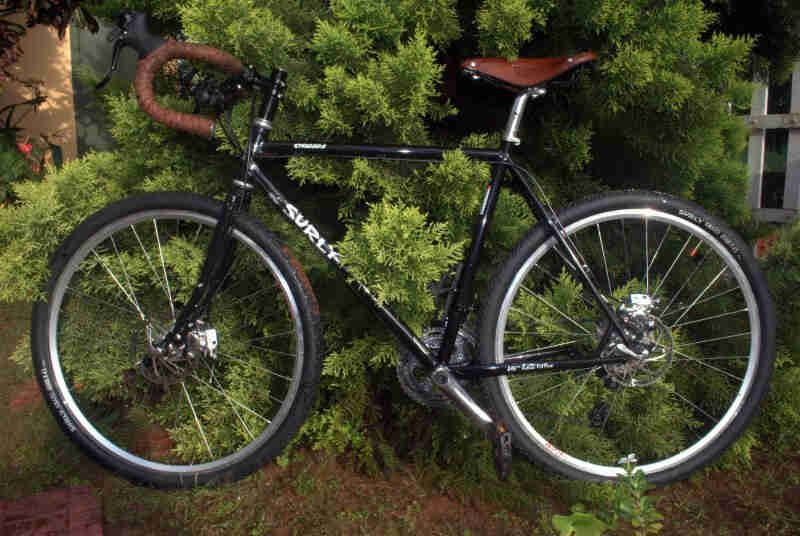 Left side view of a black Surly bike, parked against a green bush