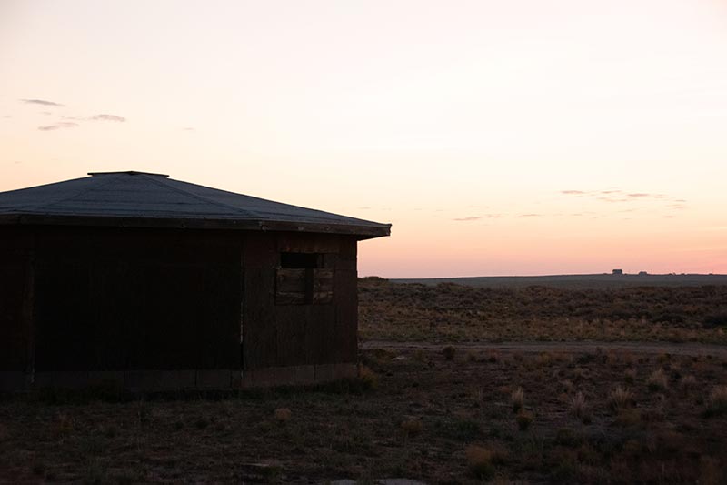View of mesa at sunset with brown yurt-style house next to gravel road.
