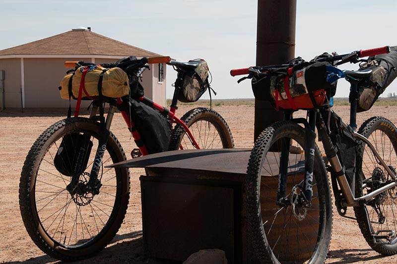 Two mountain bikes loaded with bikepacking gear parked in shade out in desert under shelter on sunny afternoon
