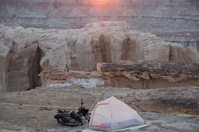 Sun setting over mesa and canyon, small tent set-up with loaded mountain bike laying on ground next to it