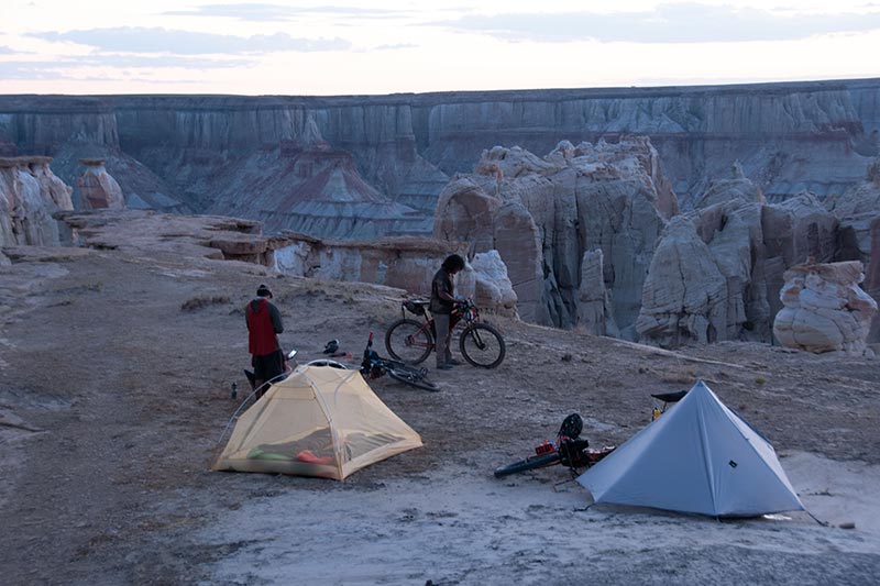 Two people getting gear from bags on their mountain bikes, tents set-up near edge of canyon post sunset