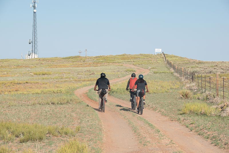 Three mountain bikers on high elevation prairie double-track road with fence on one side and radio tower in distance
