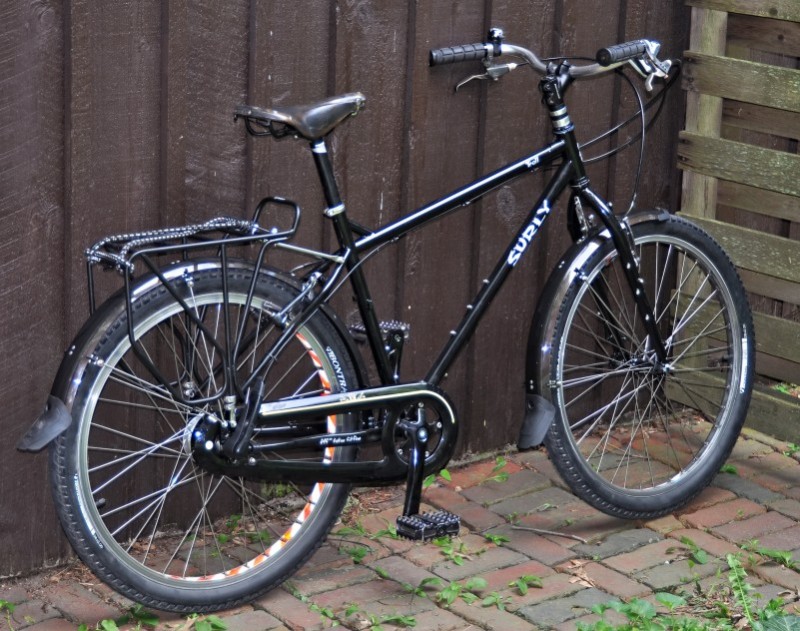 Right side view of a black Surly Troll bike, parked on a brick walkway, leaning against a wood sided wall