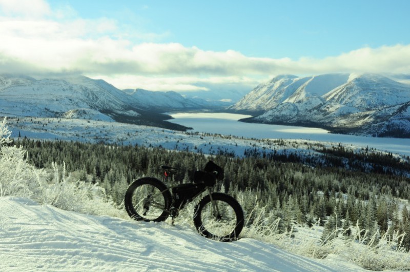 Right side view of a Surly fat bike, in a snowy field with trees, and a lake with snow covered mountains in background