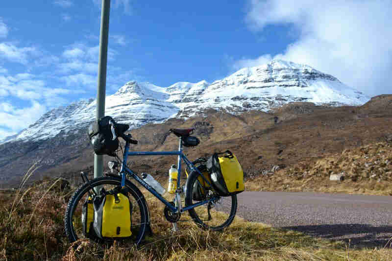 Left side view of a blue Surly bike with saddlebags, parked on a grassy roadside with snow capped mountains behind