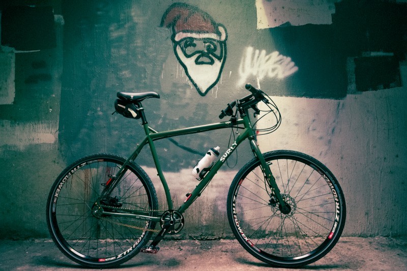 Right side view of a green Surly Ogre bike, parked next to a concrete wall with a graffiti painted Santa Claus head