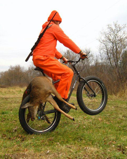Right side view of a cyclist in blaze orange clothes, riding a wheelie on a black Surly bike, with a deer laying on back