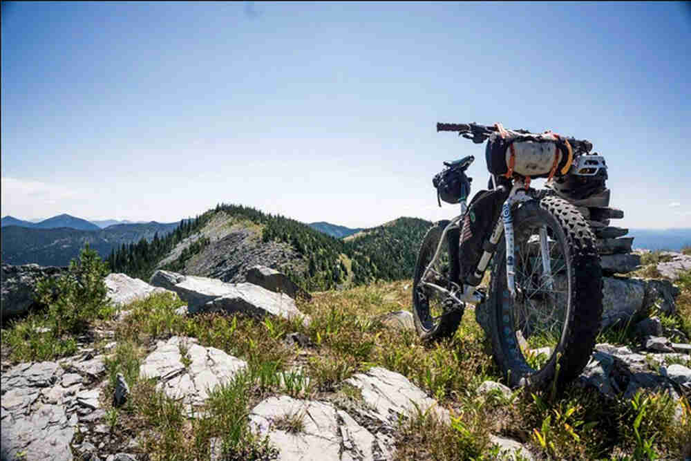 Front view of a white Surly fat bike with gear, parked on a rocky hilltop, with mountains in the background