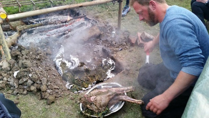 Person looking down at a pan of food in front of a food smoker pit dug into the ground