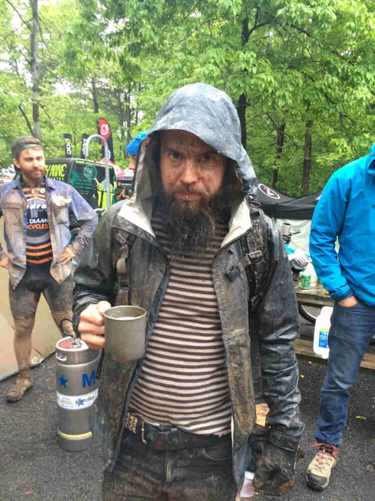 Front view of a person wear a raincoat, with a muddy face, holding a coffee cup, with people and trees in the background
