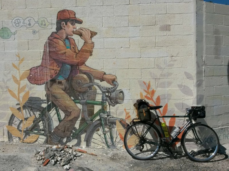 Right side view of a Surly Disc Trucker bike, leaning against a block wall with a mural of a cyclist riding a bike