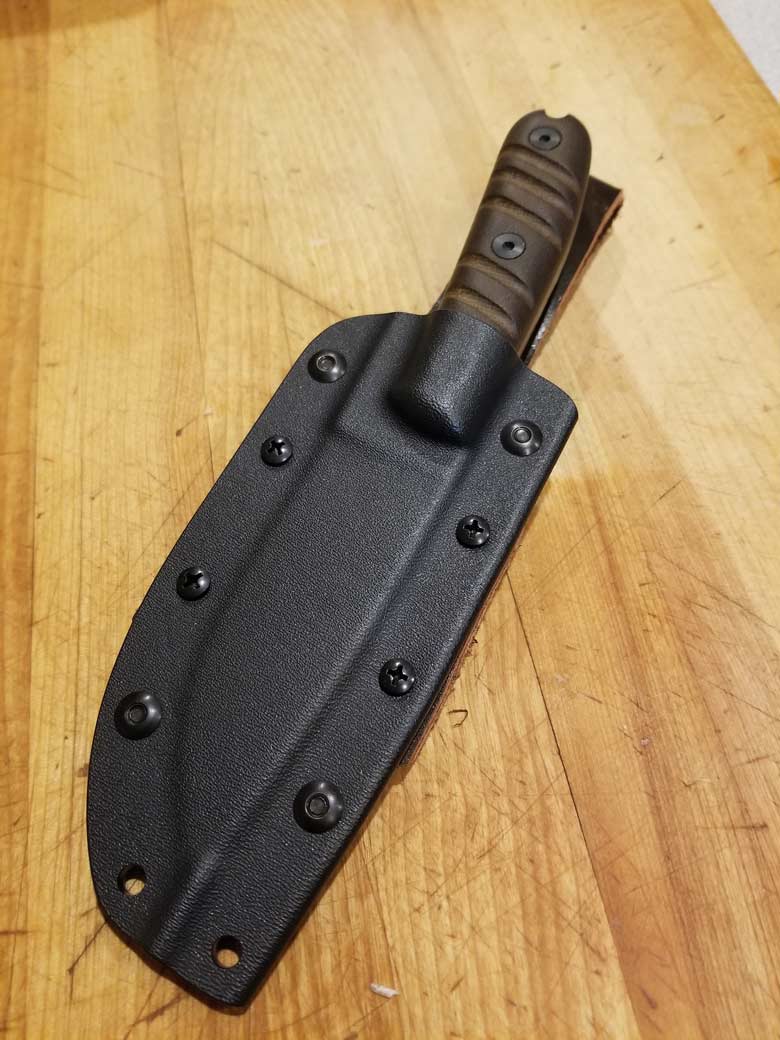 A fixed blade knife with a brown handle is inserted in a black sheath lays on a hardwood floor at an angle