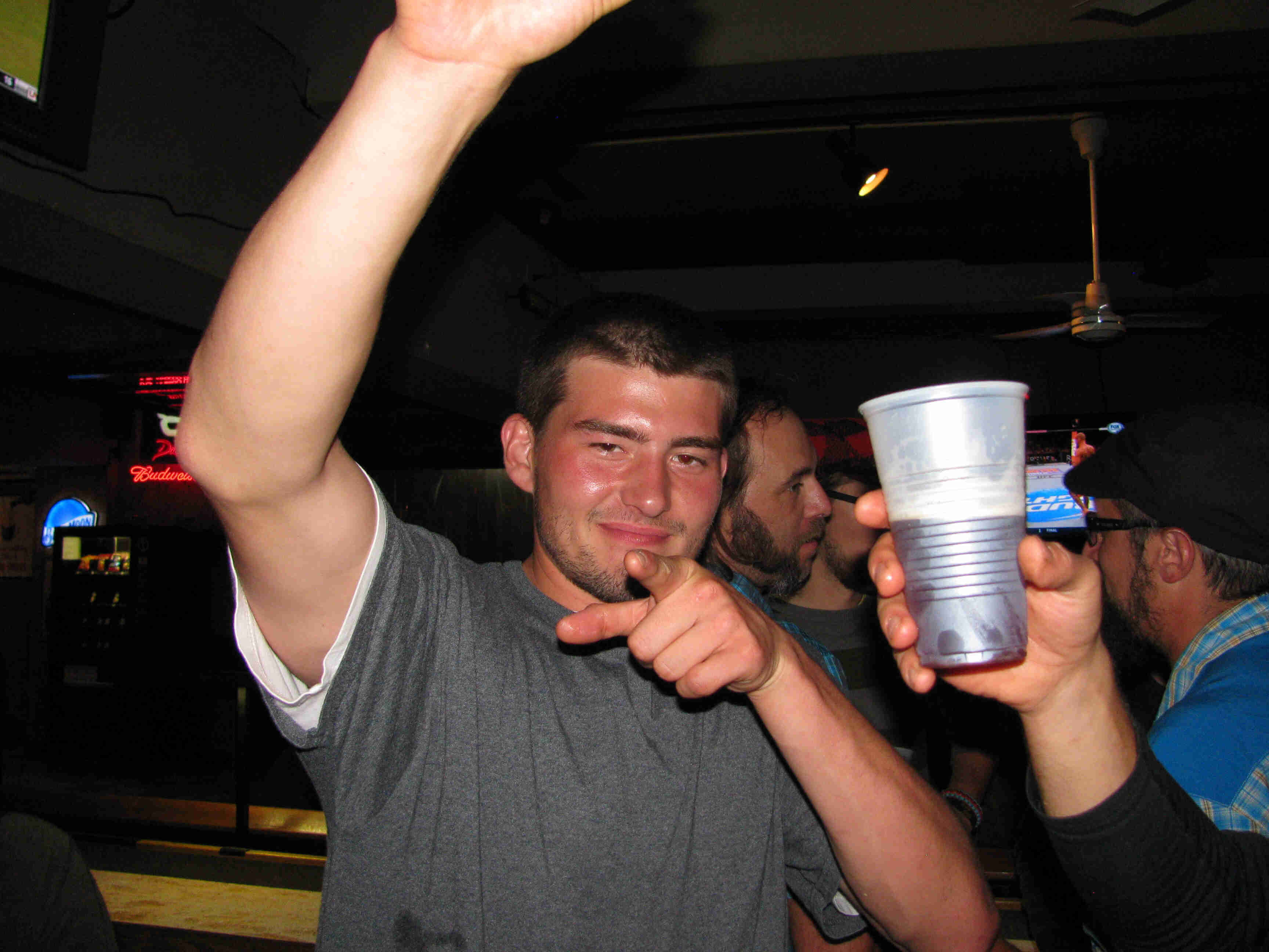 Front view of a person pointing with left their left hand and raising their right arm, in a dimly lit bar
