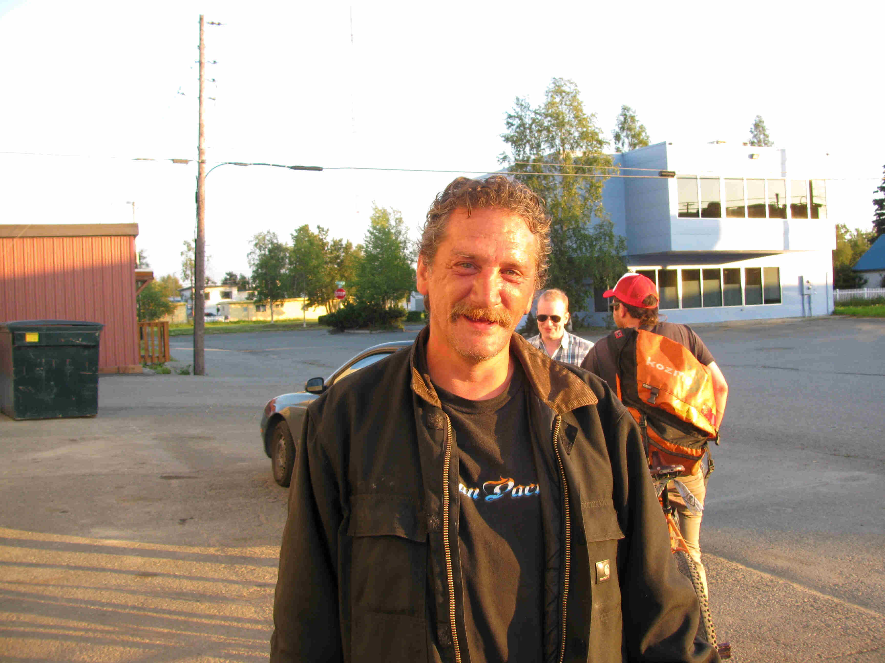Front view of a person with a mustache and wearing a black jacket, standing and posing, outside on a paved lot