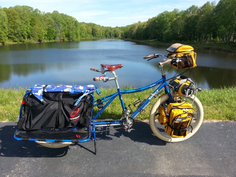 Right side view of a blue Surly Big Dummy bike with gear, parked on a trail, in front of a pond with trees all around it