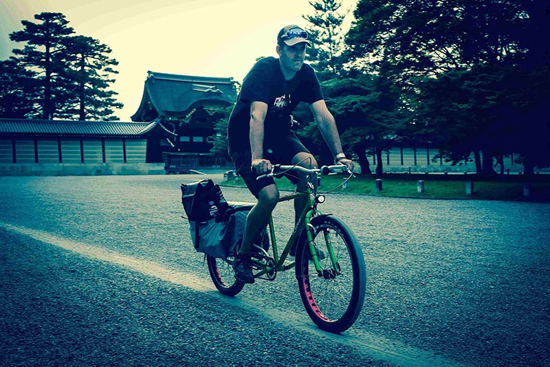 Rear, right side view of a cyclist on a green Surly Big Dummy bike in a parking lot, with a Japanese palace behind them