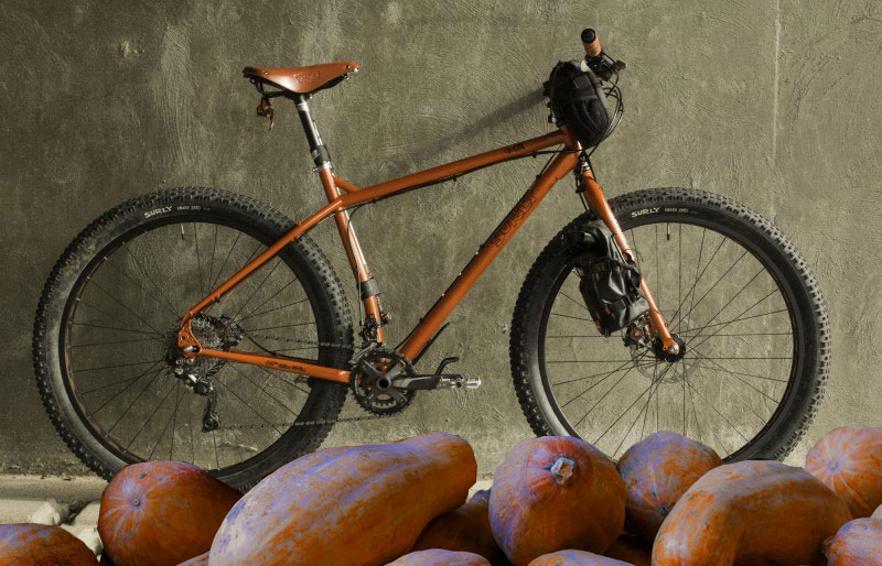 Right side of a Surly ECR bike, orange, against a cement wall, and orange colored vegetables in front of the tires