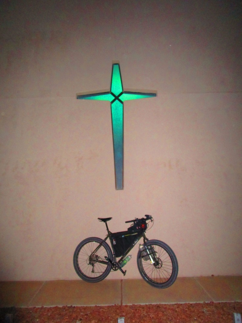 Right side view of a Surly ECR bike with frame bag, parked against a tan wall with a glowing green cross above, at night