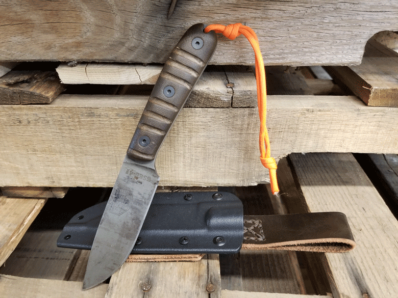 Knife with paracord tied to the wood handle leans on a stack of pallets blade face down and black sheath lying flat