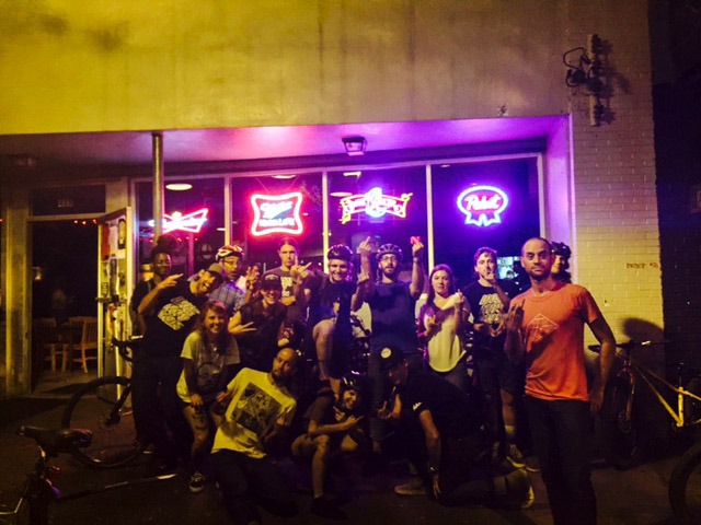 Front view of a group of cyclists, in front of a bar with neon signs at night, showing their middle fingers