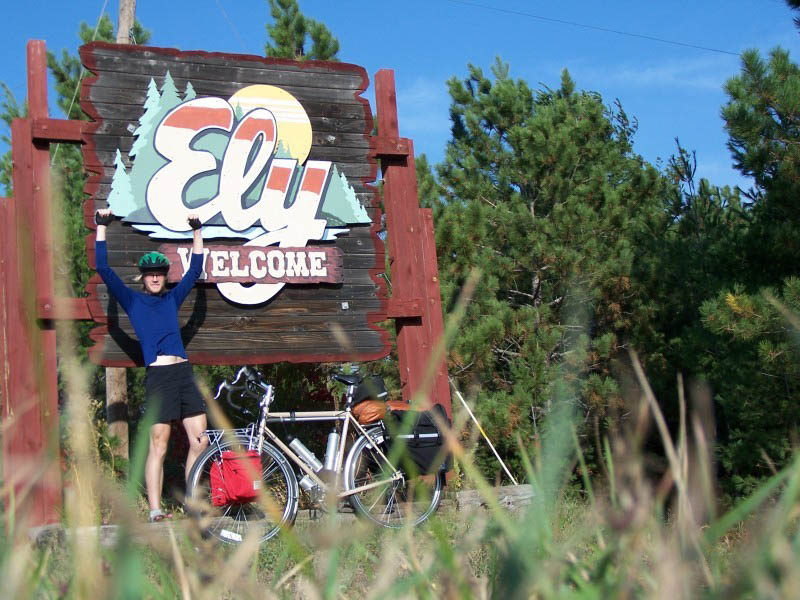 Front view of a cyclist holding up both arms, next to a bike, in front of an Ely,  welcome sign