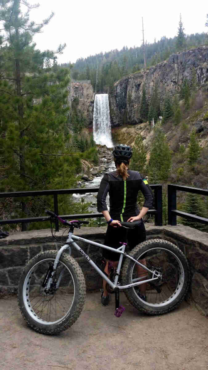 Left side view of a gray Surly fat bike, leaning on a cyclist, standing on a platform, facing a waterfall in the forest