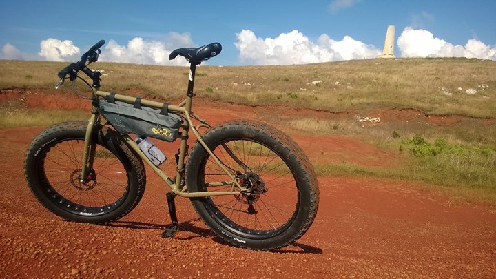 Left side view of a Surly fat bike, olive, parked on red gravel, and a grass hill with a tower on top in the background