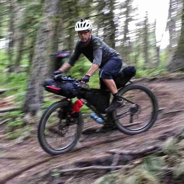 Left side view of a cyclist riding a bike loaded with gear, down a hill on a dirt trail in the forest