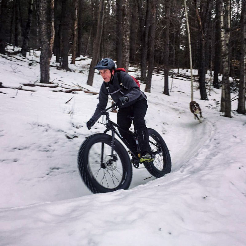 Front view of a cyclist riding a Surly fat bike down a hill, on a snowy trail, with a dog chasing behind