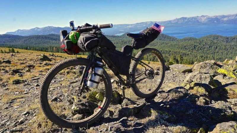 Left side view of a Surly bike with gear, parked on a rocky hilltop, with a forest and mountains in the background