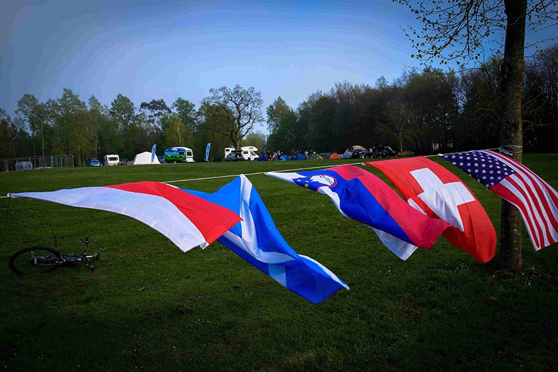 Side view of a rope, tied to a tree with the flags of countries hanging from it, and a field with a campsite behind