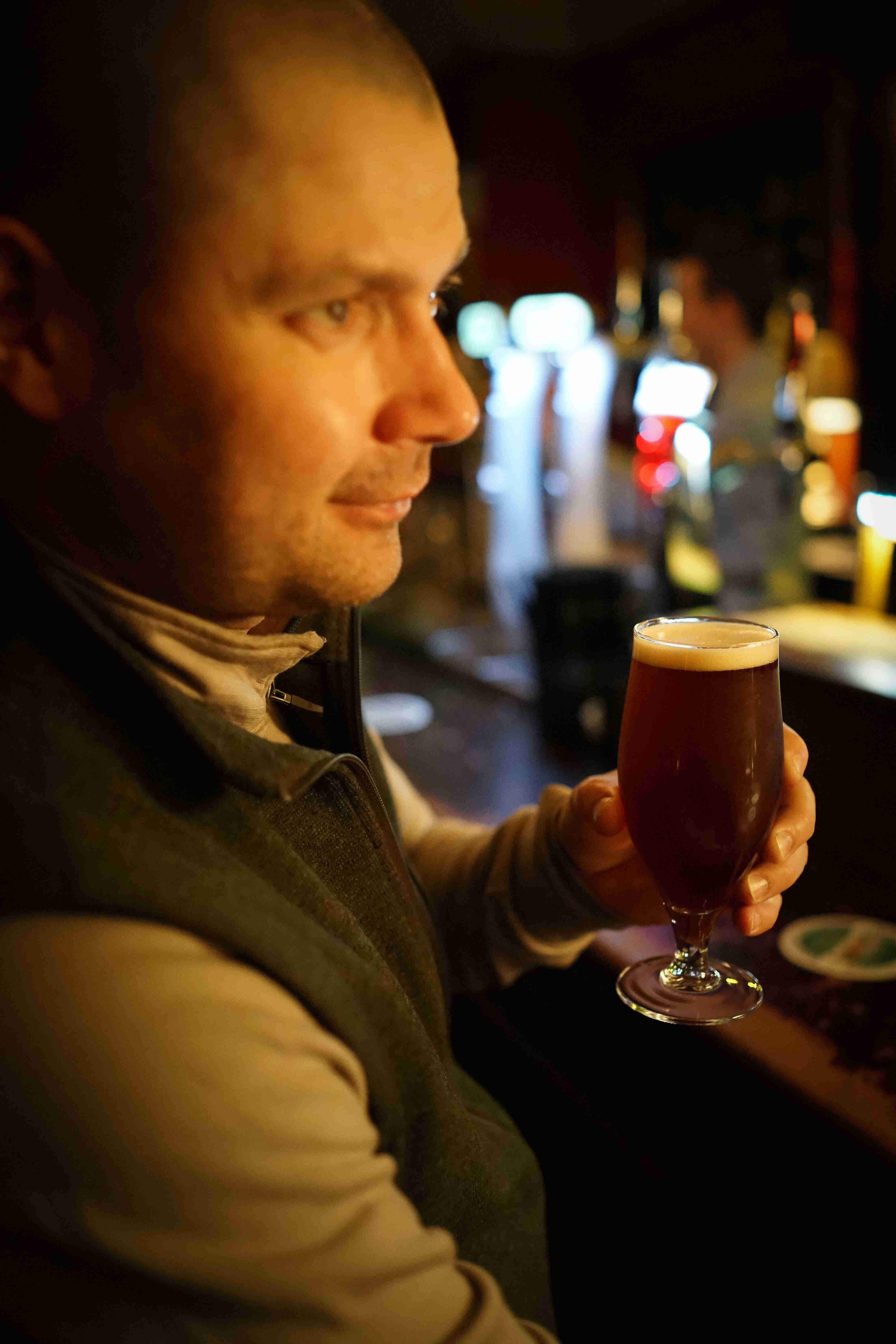 Headshot of the right side of person, holding a glass of beer, inside of a bar