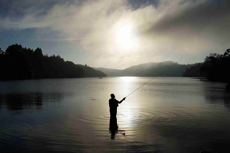 Silhouette of a person standing in a lake, while fly fishing during a sunrise, with trees and hills in the background