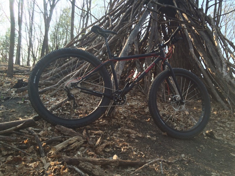 Right side view of a black Surly Krampus bike, parked on dirt hillside, in front of a teepee shaped pile of branches