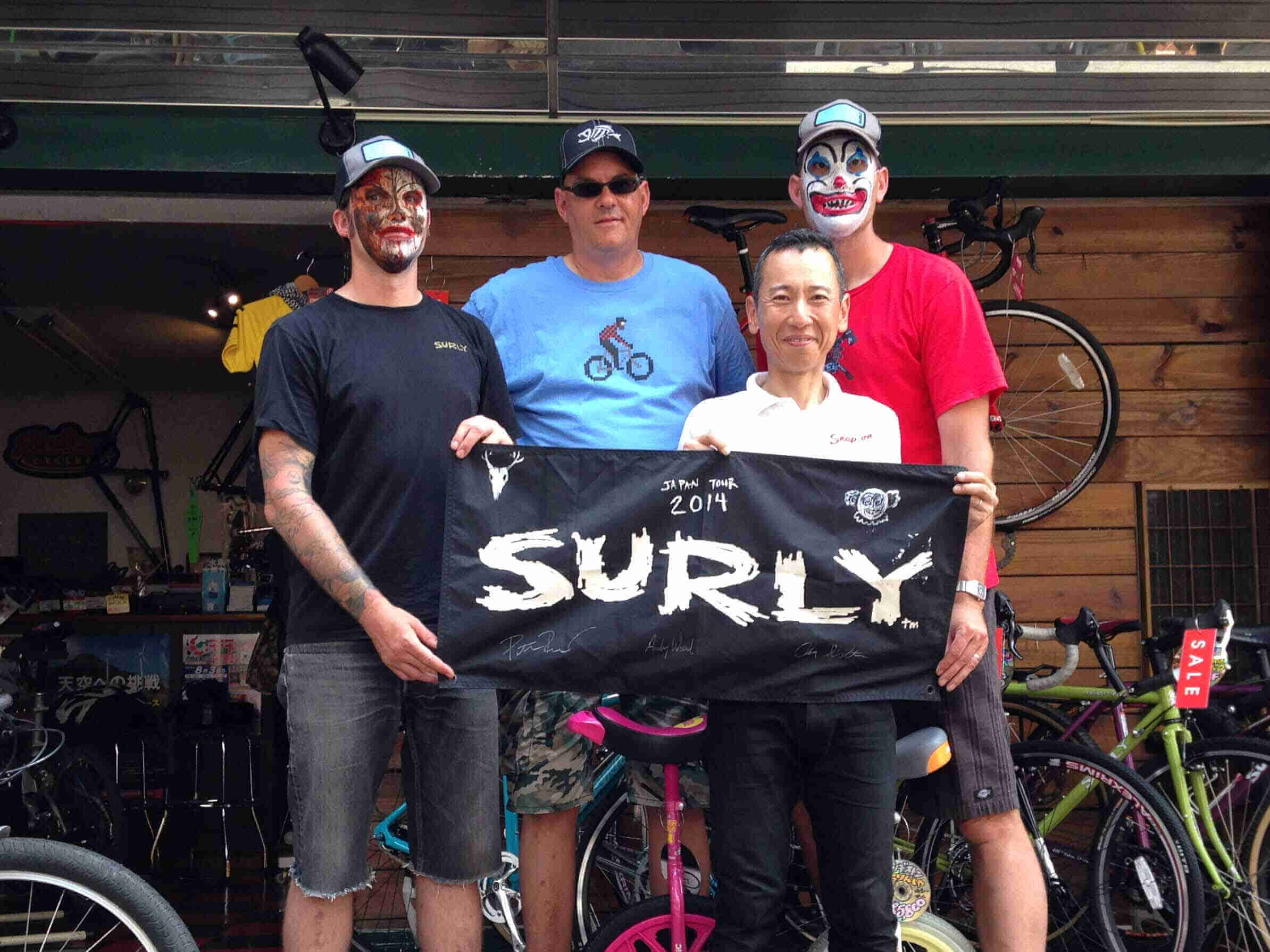 Front view of 4 people posing with a Surly banner in front of them, with a bike shop behind in the background