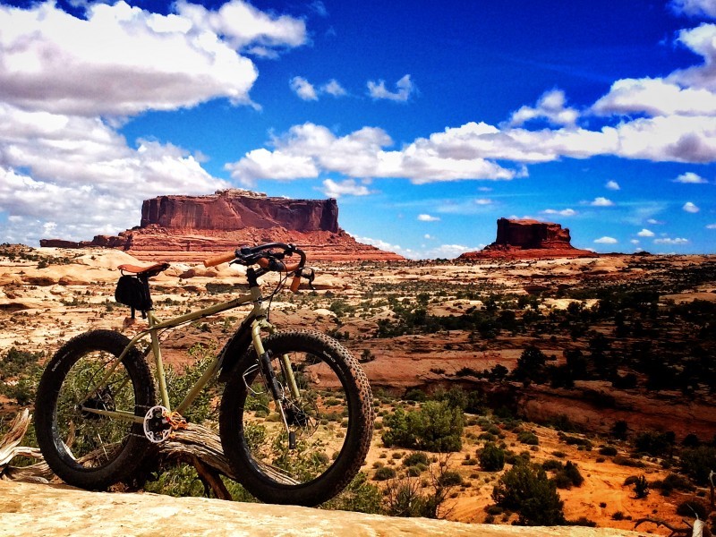 Right side view of a gold Surly fat bike, with a hilly, bushy desert and red buttes in the background