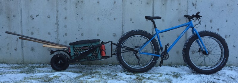 Right side view of a blue Surly fat bike and trailer packed with a crate and boards, leaning against a cement wall