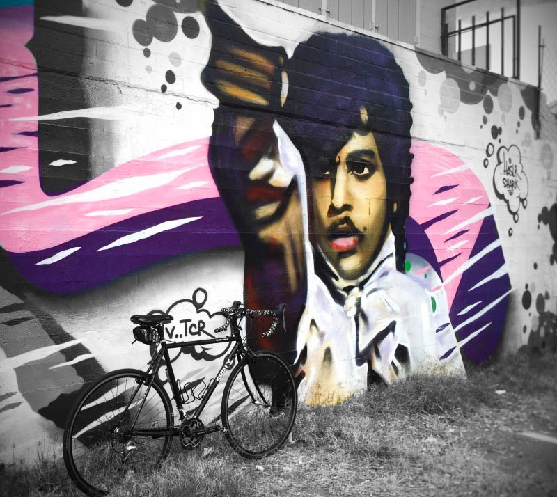 Right side view of a black Surly bike, standing in grass, leaning on a wall with a mural of Prince, the musician