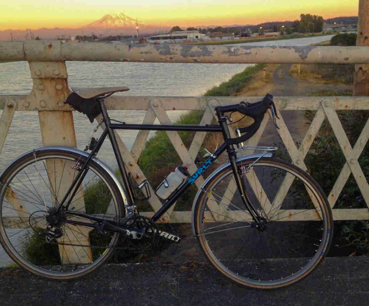 Right side view of a black Surly bike against the rail on a river bridge, with a snow capped mountain in the background