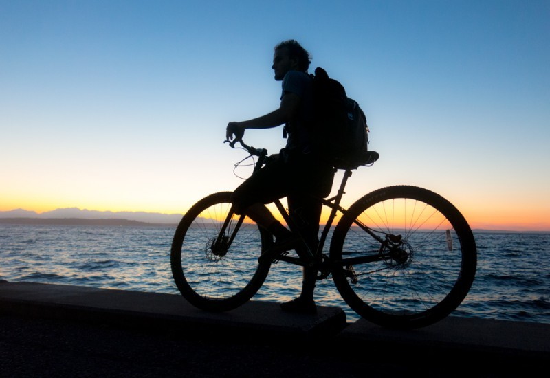 Dimly lit, left side view of a cyclist, standing over a Surly bike, on a wall with the ocean behind, at sunset