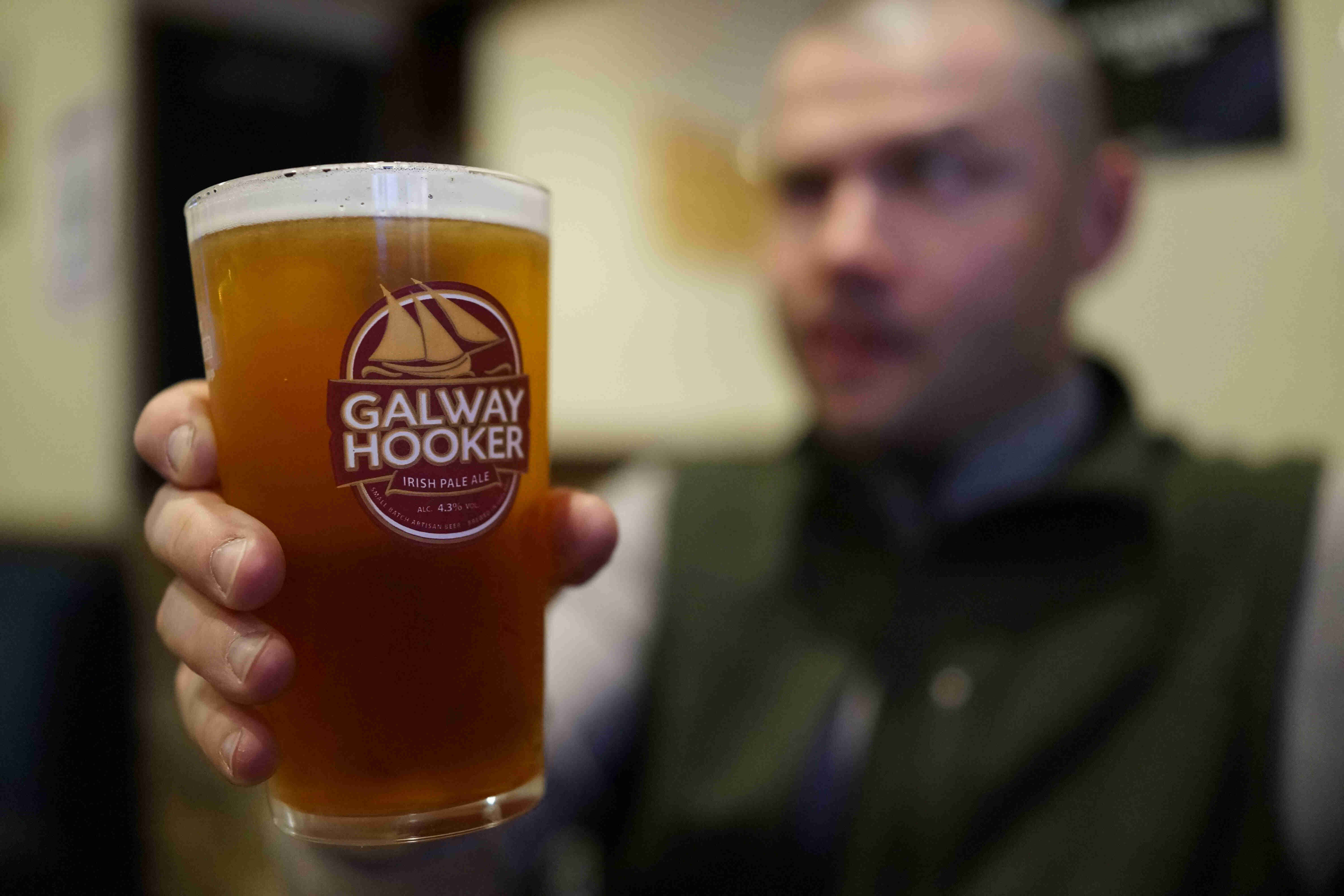 Close up of a glass of beer with a Galway Hooker logo on it, with a blurred person holding it up from behind
