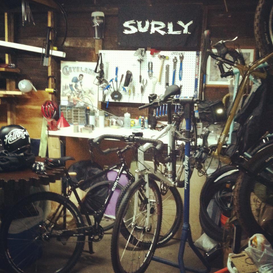 Various Surly bikes, in front of a workshop bench, with a Surly banner above a pegboard with tools