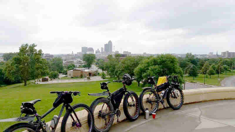 Three bikes with gear, parked in line against a short cement wall, with a green field and city skyline in the background