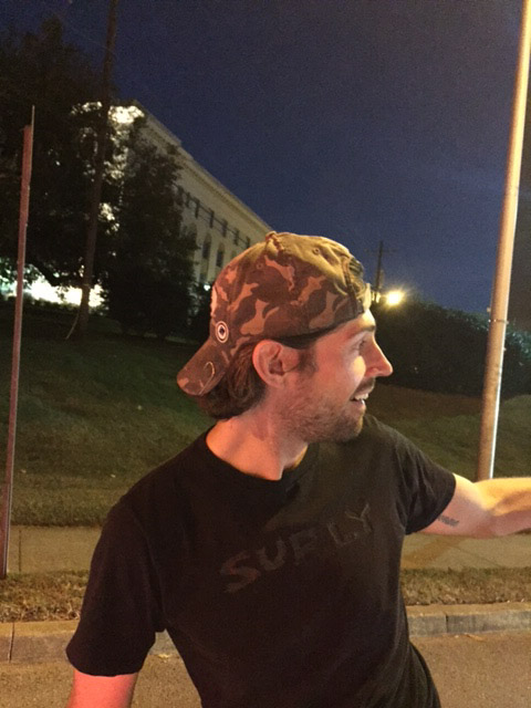 Right profile of a person wearing a camo baseball cap backwards, on a street at night