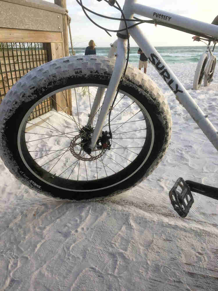 Cropped, front left side view of a white Surly Pugsley fat bike, parked in white sand,  with the ocean in the background