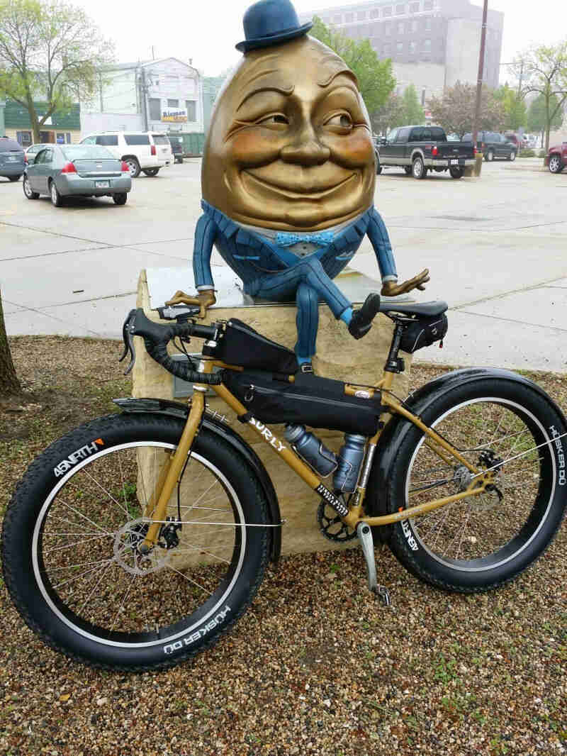 Left side view of a tan/brown Surly bike, parked against a humpty dumpty statue, with a parking lot in the background
