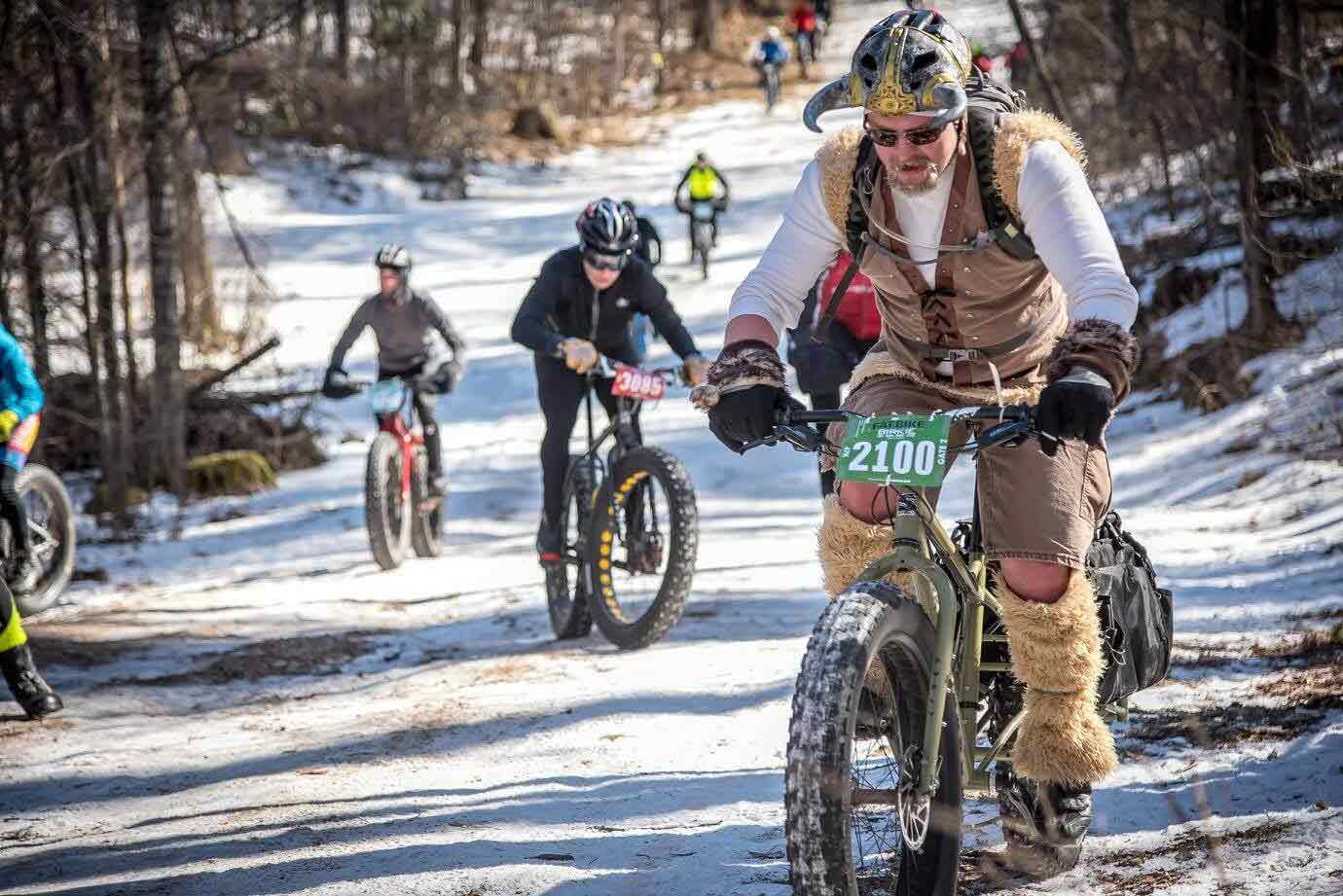 Cyclists ride up an icy road on fat bikes  in the woods with front cyclist wearing viking clothing