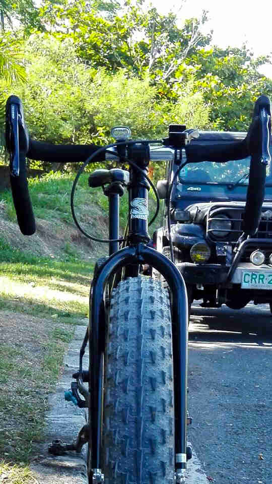 Front view of a black Surly Ogre bike with a front fat tire, with a jeep and trees in the background