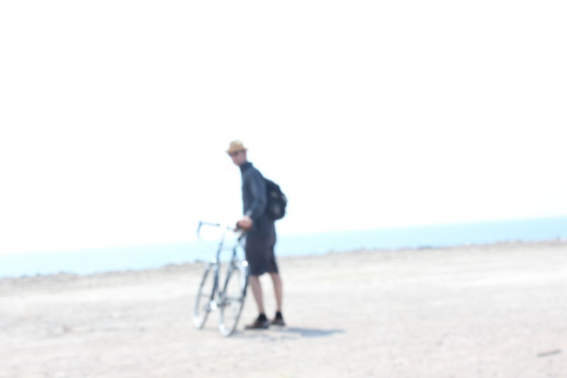 Hazy, rear view of a cyclist looking back, while standing on the right side of a bike, on a sandy lot with ocean behind